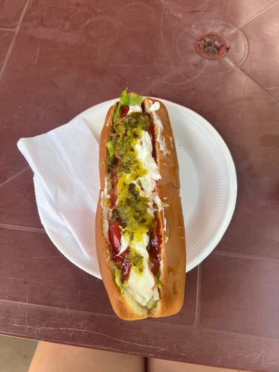 A single, loaded hot dog is presented on a white paper plate next to a stack of white paper napkins. The hot dog is in a bun and topped with ketchup, mayonnaise, relish, and lettuce. The plate rests on a brown, textured table with a circular indentation in the center. The scene is photographed from a top-down perspective, with a person's legs visible at the bottom of the frame. This image was taken on the street in the city of León, Nicaragua.