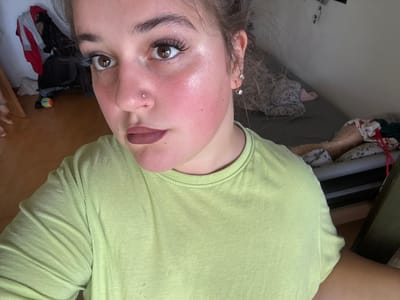 A young woman with brown hair pulled back is looking into the camera, with her face taking up most of the frame. She is wearing a light green t-shirt. She has on heavy eye makeup, including false eyelashes, and a matte brown lipstick. She has a small silver stud piercing in her nose. Her cheeks are flushed pink and appear to have a dewy, highlighted finish. She is indoors, in what appears to be a bedroom, with a bed and clothing visible in the background. There is a wooden floor in the foreground.