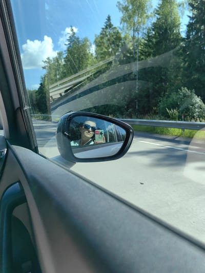 A selfie is taken by a woman from inside a moving vehicle, reflected in the side-view mirror. She is wearing sunglasses and a light blue top, holding a smartphone in her right hand. The background visible through the car window and in the mirror shows a road lined with trees and a clear blue sky with scattered clouds, suggesting a rural or suburban setting. The scene appears to be a daytime drive on a paved road, possibly a highway, given the presence of a guardrail. The reflection indicates the woman is the passenger, as the driver is not visible in the mirror's reflection. The context of Ракаўскі сельскі Савет in Belarus is not directly evident from visual cues within the image itself, but the description aligns with a typical road trip scenario that could occur in such a location.