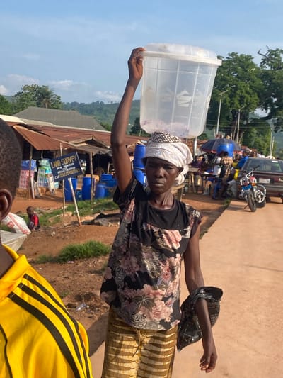 This detailed image, set in Amaseri, Nigeria, captures an outdoor street scene under clear daylight. In the foreground, an adult woman with dark skin stands facing slightly left, carrying a large, translucent ribbed plastic container with a white lid balanced on her head, stabilized by her raised right hand. She wears a white patterned headwrap, a black short-sleeved top with a floral print featuring pink and white elements, and light brown horizontally striped pants. Her left arm is bent, holding a dark plastic bag. To her immediate left, the back of another person is partially visible, wearing a yellow garment with three black stripes on the shoulder.
The mid-ground reveals a reddish-brown dirt ground interspersed with patches of green grass. A blue sign mounted on white poles stands prominently to the left of the central woman, displaying "LAND SURVEY" at the top, followed by "NEW NUMBER AND REGISTRATION," and a contact number "Call 08035492559." Behind this sign and extending further left are several rudimentary structures with corrugated metal roofs, suggesting market stalls or small shops. Numerous blue plastic barrels are visible near these structures. A small child in a red shirt is seated on the ground to the left of the sign.
To the right of the central woman, a paved surface delineates a walkway or road where a red and chrome motorcycle is parked, facing left. Behind it, a dark-colored sedan is partially visible. Further back, several indistinct figures are gathered under a large dark umbrella, indicative of a marketplace or social gathering. The background features lush green trees and distant hills under a clear blue sky with minimal white clouds.