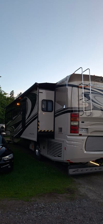 A fleetwood Revolution RV is parked on a grassy area next to a gravel path. The RV is light beige with a dark gray and black graphic design. The driver's side of the RV is visible, with the awning extended. A silver ladder is attached to the rear of the RV. A black car is parked to the left of the RV. Trees are visible in the background. The sky is clear and pale blue. The license plate on the RV reads MOB-763. The RV has a "REVOLUTION" logo on the rear. The image is taken from a low angle.
