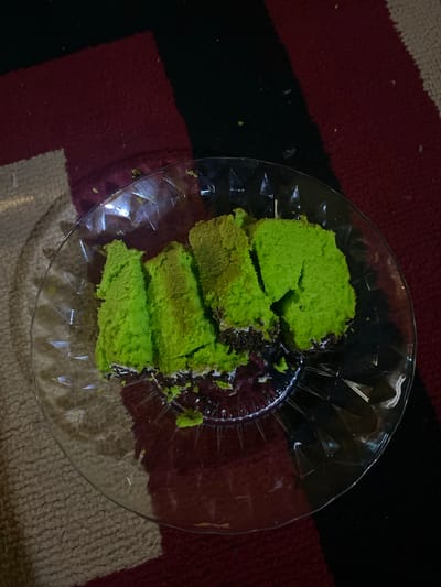 In Tanjongkapai, Indonesia, a clear, circular glass plate rests on a multi-colored patterned floor covering. The plate contains four irregularly cut pieces of bright green cake. The cake pieces exhibit a soft, spongy texture. Along the bottom edge of some cake sections, a white layer, possibly frosting or cream, is visible, topped with small, dark brown cylindrical sprinkles or chocolate shavings. Minor crumbs of green cake and a few dark sprinkles are scattered on the glass surface. The floor covering features distinct blocks of color: dark red, black, and a light beige or off-white, suggesting a rug or carpet. The lighting is moderate, creating reflections on the glass plate. No individuals or actions are present.