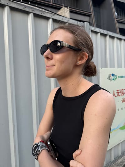 An outdoor, slightly rotated portrait captures a person, likely female, with light skin and brown hair tied in a bun. She is wearing black sunglasses and a black sleeveless top. On her left wrist, she wears an intricate watch featuring a dark face and a silver wire-wrapped design, with a red string or band integrated into it. She holds a smartphone in her left hand, which is tucked close to her body. A small, silver stud earring is visible in her left ear.

The setting appears to be a brightly lit outdoor or semi-enclosed area, suggesting daytime. To the left, dark, reflective panels are visible, possibly windows or building elements. The lower part of the frame shows horizontal ridged surfaces, which could be corrugated metal walls or steps. In the background, behind the person's right shoulder, a light-colored sign features Chinese characters and a green leaf logo with blue accents. Part of the text reads "人无信不..." (Rén wú xìn bù...), which translates to "If people have no trustworthiness/credibility, they cannot..." or "Without trust, people cannot..."

The person is looking downwards and to her left, with a neutral expression, possibly a slight smile, obscured by her sunglasses. The image was captured in Putuo District, China, consistent with the visible Chinese text.