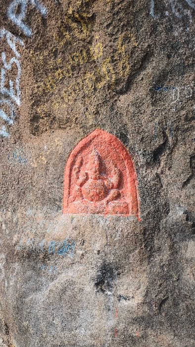 A reddish-orange relief carving of the Hindu deity Ganesha, depicted seated with an elephant head and multiple arms within an arched recess, is integrated into a rough, weathered rock or concrete surface in Kadaladi, India. The surrounding surface exhibits a granular texture with varied coloration, including tones of grey, brown, beige, and yellow. Graffiti in white and yellow pigments is present in the upper left and upper right sections, while faint blue markings are visible on the lower left. Darker smudges and streaks are observable across the surface, particularly below the carving. No individuals are present in the scene. Ambient light illuminates the subject, emphasizing the relief of the carving and the texture of the rock.