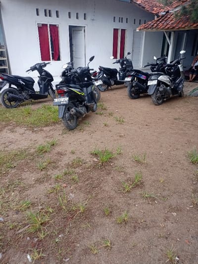 The image shows six motorcycles parked on a dirt ground in front of a white house. There are windows with red curtains on the house. There are several plants growing on the ground. The motorcycles are all black and have license plates.  The scene is located in Pameungpeuk, Indonesia, at coordinates -7.668 latitude and 107.694 longitude.