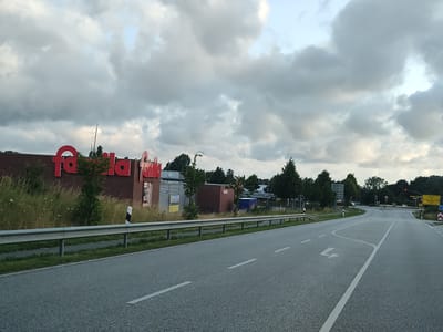The image depicts a road scene in Heiligenhafen, Germany. On the left side of the road, there is a brick building with large red lettering that appears to spell "famila". Adjacent to the building is a metal guardrail bordering a grassy area with tall weeds and scattered wildflowers. The road itself is asphalt with white lane markings, including a turn arrow. Further down the road, there are trees and traffic signs. The sky is overcast with dense clouds. No people are visible in the image.
