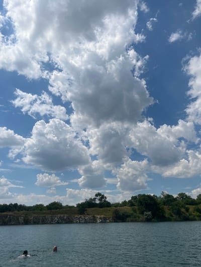 The image depicts a serene natural scene with a body of water on the right side and a sky filled with cumulus clouds on the left. The water appears calm with gentle ripples reflecting the light. Along the edge of the water is a rocky embankment covered in lush green trees and vegetation, suggesting a terrestrial boundary.

In the water, two people are visible, partially submerged. One person, closer to the foreground, appears to be swimming or wading, with their head and shoulders visible. Another person is further in the distance, with only their head visible above the water.

The sky is a vibrant blue, interspersed with numerous fluffy white clouds of varying sizes and densities, creating a dynamic visual contrast. The sunlight suggests it's daytime, likely late morning or afternoon, given the brightness and the shadows cast by the clouds.

The scene appears to be a recreational area, possibly a quarry or a natural lake, given the rocky edges and clear water. The overall mood is peaceful and relaxing, evoking a sense of enjoying nature on a pleasant day. There is no visible text in the image. The location is identified as Novomykolaivka, Ukraine.