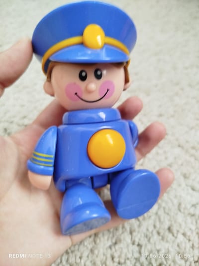A close-up, high-angle shot captures a blue plastic toy figure resembling a pilot or officer being held in a person's palm against a fuzzy, beige carpeted surface. The figure is predominantly blue with yellow accents on its cap and a yellow circular emblem on its chest. It has a pink-cheeked face with black eyes and a smiling mouth. The toy's arms are bent, and its legs are positioned as if sitting. The person's thumb is visible on the left side of the frame, gently supporting the toy. The background is a soft-focus carpet, creating a shallow depth of field that emphasizes the toy. The image contains text overlays: "REDMI NOTE 13" in the bottom left corner and "19.06.2026 10:56" in the bottom right corner, indicating the device and date/time of capture. The location of Асан-Чек, Kyrgyzstan is not visually present in the image.