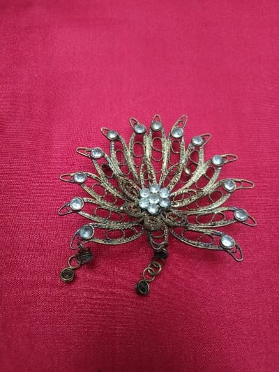 A single ornate metal brooch is centrally displayed on a deep red fabric surface. The brooch, constructed from dull gold-toned or bronze-toned filigree metal, features a multi-petal or radial design. A central cluster of seven clear, faceted stones is surrounded by eleven individual clear, faceted stones, each set within an elongated, openwork petal. Two small, coiled metal elements with dark spherical ends extend from the bottom of the brooch. The background is a plain, solid deep red textile with a fine weave and slight sheen. No individuals, actions, or interactions are visible. The scene is situated in Horní Vltavice, Czechia.