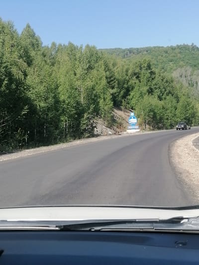 The image captures a paved road winding through a forested mountainous landscape under a clear blue sky. On the left side of the road, a dense forest of deciduous trees with green foliage lines the embankment. On the right side, the road curves, with a gravelly shoulder and more trees extending up the hillside. In the distance, a dark-colored SUV is visible driving on the road. Further down the road, a sign with blue and white graphics, possibly indicating a location or attraction, stands on a concrete base. The foreground shows the interior of a vehicle, including the windshield and dashboard. The environment suggests a rural or natural setting, possibly a scenic route within or near the city of Ushanovo, Kazakhstan.