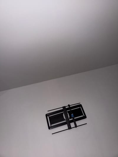 A black metal TV wall mount is affixed to a white wall. The mount is folded inwards, with its arms extending outwards. The scene is indoors, with a white ceiling visible in the upper portion of the frame. The lighting is neutral. The image was taken in Vernon Hills, United States of America.
