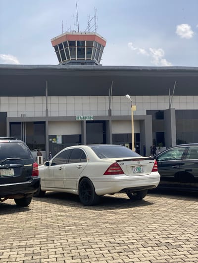 The image depicts an outdoor scene at an airport, likely in Kwi, Nigeria, focusing on the departure terminal, an air traffic control tower, and parked vehicles. The ground is paved with interlocking concrete blocks.

The main building is a modern airport terminal featuring a facade of gray panels and glass. Above one of the entrances, the word "DEPARTURES" is prominently displayed in green letters. To the left of the terminal stands a distinctive air traffic control tower with an octagonal, glass-enclosed cab and numerous communication antennas extending from its structure.

In the foreground and midground, several vehicles are present. A white Mercedes-Benz C32 AMG sedan is parked with its rear facing the viewer. Its Nigerian license plate, "NSR-36TEA," from Lagos State, is clearly visible, as are the "C32" and "AMG" badges on the trunk. Behind the Mercedes, the rear of a black Toyota Highlander SUV is partially visible, also bearing a Nigerian license plate starting with "LAGOS." Another dark-colored car is partially visible on the far right.

A few individuals are discernible near the terminal entrance and between the parked cars, indicating people moving around the airport. The sky is bright blue with scattered white clouds. The presence of clear, relatively short shadows suggests it is a sunny day, likely late morning or early afternoon. A street light pole is also visible near the terminal entrance.