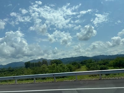 The image captures a scenic view from a roadside, likely along a highway or main road, in or near the municipality of Chibati, Georgia. The foreground is dominated by an asphalt road with a white lane marker. A metallic guardrail runs horizontally across the middle ground, separating the road from the landscape beyond. This guardrail is supported by concrete posts.

Behind the guardrail, a lush green landscape unfolds, characterized by rolling hills covered in dense vegetation and trees. In the distance, a range of mountains forms the backdrop, their peaks softened by the atmospheric haze. The sky above is a vibrant blue, dotted with an array of fluffy white cumulus clouds, some of which cast shadows on the landscape below. The overall impression is one of a bright, sunny day in a rural, mountainous region. There are no people or specific structures visible in the image.