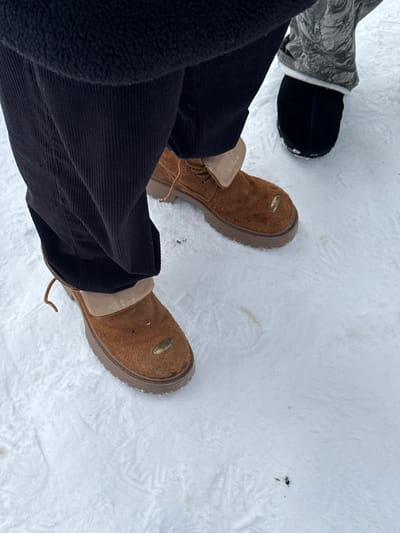 The scene is set outdoors in Yeghipatrush, Armenia, on a ground uniformly covered with textured, compacted snow that contains minimal dark particulate matter. Two individuals are present, visible from the lower torso downward. The subject in the foreground is wearing black corduroy pants and brown, mid-cut, suede-like boots with thick, light brown lug soles. The boots feature tan fabric ankle cuffs and visible lacing, with one loose lace on the left boot. A small, oval, metallic gold-colored tag is affixed to the toe area of each boot. Faint white alphanumeric text, including "G-LONA," "(5ZJ) П-20," and "П3-12-ДР," is visible on the inner ankle cuff of the right boot. The second individual, positioned partially behind and to the right of the foreground subject, is wearing grey, possibly camouflage-patterned or textured pants and black slip-on footwear. Both subjects are standing on the snow.