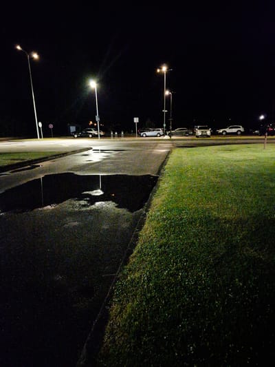 A nighttime image depicts a wet asphalt surface in the foreground, with a large puddle reflecting light from nearby streetlights and vehicles. The puddle is situated at the edge of a grassy area. In the background, several cars are parked or driving on a road, illuminated by streetlights. The scene appears to be a parking lot or roadside area in Pabradė, Lithuania.