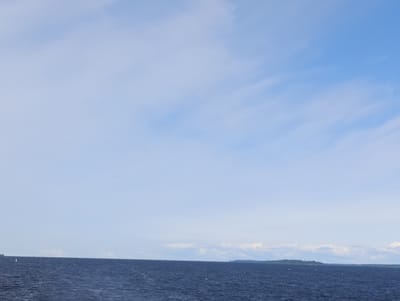 This media file depicts a serene seascape with a vast expanse of the Baltic Sea under a pale blue sky dotted with wispy clouds. The horizon is clearly defined, with a sliver of land visible in the distance, suggesting a coastal or island setting. On this distant landmass, a small lighthouse or tower can be discerned, hinting at a navigational landmark. The water itself is a deep, dark blue, rippled by a gentle breeze, reflecting the muted tones of the sky. A lone bird, likely a seagull, is captured in flight, adding a touch of life to the otherwise tranquil scene. The overall impression is one of peace and openness, likely captured during daylight hours, given the brightness of the sky. The location context of Oulu, Finland, suggests this could be a view from the coast or a vessel on the Gulf of Bothnia. There is no visible text in the image.