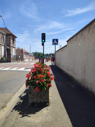 The image depicts a street scene in Ressons-sur-Matz, France, on a clear day with a blue sky and scattered clouds. In the foreground, a large concrete planter filled with vibrant red flowers sits on the sidewalk. The planter casts a long shadow across the asphalt. To the left of the planter, a pedestrian crossing with white triangular markings on a red background is visible across the street. Adjacent to the crossing, a traffic light shows a green signal.
Further down the street on the left, a two-story building with a brick facade and red-tiled roof is visible. A black metal fence borders the property. On the right side of the street, a textured, light-colored wall runs alongside the sidewalk. A blue road sign with a white upward-pointing triangle inside a white triangle is mounted on a pole in front of the wall. Standing on the sidewalk near this sign is a life-sized figure of a person wearing a red jacket, white pants, a red hat, and a black backpack. The figure appears to be a mannequin or a statue. Another traffic light further down the street also shows a green light. The street is paved with asphalt, and there is a curb separating the sidewalk from the road. The environment appears to be a quiet, residential or small-town street.