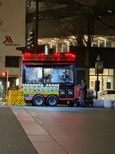 A brightly lit Halal food cart is situated on a city sidewalk at night. It boasts "Staple of Charlotte Halal Food Cart" in prominent lettering. A customer in a coat is engaging near the cart, which has menu pictures and "Halal Food Cart" displayed. The background shows a "Marriott" building, a "Green" sign, bare tree branches, and illuminated building windows. Yellow safety barriers are positioned near the cart.