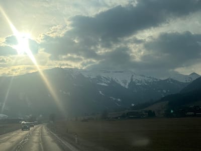 The image displays a panoramic view of a mountainous landscape in Steindorf, Austria, under a partly cloudy sky with prominent sunlight. In the foreground, a wet asphalt road curves from the lower left towards the mid-left, occupied by two vehicles traveling away from the viewer. The lead vehicle is a light-colored van or SUV with active headlights, followed by a darker vehicle. White and red road markers are visible along the right edge of the road. To the right of the road, a large expanse of fallow or harvested brown field extends towards a cluster of traditional buildings, indicative of a village or hamlet, nestled at the base of the mountains.

The background is dominated by snow-capped mountains, with dark green coniferous forests covering their lower slopes. A prominent mountain peak in the left-center features two distinct structures, likely communication towers or antennae, on its summit. The sun, positioned in the upper left, generates significant lens flare, partially obscured by dense, dark-grey clouds that cover much of the sky, particularly in the upper right quadrant. The overall lighting suggests either early morning or late afternoon, with the sun breaking through the cloud cover.