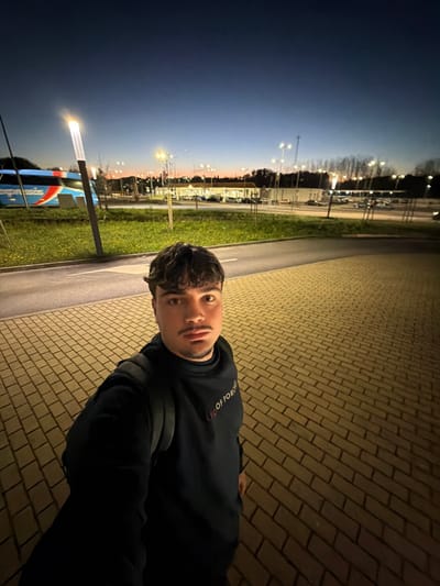 The image depicts a male individual standing on a brick-patterned paved surface in Penafiel, Portugal, during twilight. The sky transitions from dark blue overhead to an orange-pink glow on the horizon. Artificial lighting from numerous lampposts illuminates the scene.
The man, positioned centrally and slightly to the left, is taking a selfie, holding the camera at arm's length. He has short, curly dark brown hair, a light mustache, and a short goatee. He is wearing a dark, possibly black or navy blue, long-sleeved hooded sweatshirt with a white, red, and yellow graphic on the chest, partially visible as "OF POR." He also wears a dark backpack.
Behind the individual, a narrow asphalt road or bus lane runs horizontally. To the left of the road, a grassy median strip features a single, illuminated lamppost with a rectangular light fixture, and some small plants. A large blue and white "EXPRESSOS" branded bus is parked on this side of the road.
Further in the background, a sprawling paved area, likely a parking lot, is visible, containing multiple parked vehicles and illuminated by numerous tall lampposts emitting a yellowish light. Beyond this area, commercial-style buildings and a distant treeline are discernable. The overall environment suggests a bus terminal or a large commercial complex with significant parking infrastructure.