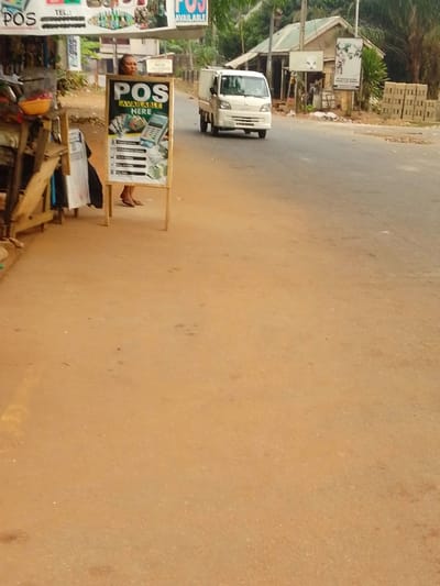 This image depicts a roadside scene in Umuoji, Nigeria. In the foreground, on the left, a wooden market stall with a yellow bowl containing red items is visible, partially obscured. A woman, dressed in a dark top, stands behind a prominent A-frame signboard in the midground. The signboard, black with yellow text, reads "POS AVAILABLE HERE" and displays images of a point-of-sale (POS) machine, currency, and a mobile phone, alongside listed services such as "Cash Withdrawal" and "Bill Payment." The woman's head, shoulders, and sandaled feet are visible. To the right, a white commercial van travels on a dark asphalt road. The road shoulder is reddish-brown earth. In the background, several single-story buildings with light walls and dark roofs are visible, interspersed with green foliage. A stack of grey concrete blocks is present on the far right. Multiple advertising signs are affixed to the buildings, including one with a faint, indistinct image.