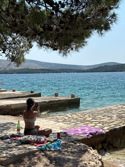 A woman is sitting on a stone ledge by the water, taking a selfie with her phone. She is wearing a bikini. Scattered around her are towels, flip-flops, and a bottle of water. In the background, the blue water stretches out to a tree-covered coastline under a clear sky. The scene is likely a beach or a waterfront promenade within the city of Šibenik, Croatia.