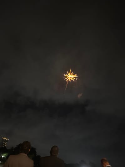 An outdoor nighttime shot shows a single yellow firework exploding in a cloudy, dark sky. In the foreground, three people are visible from the back, observing the display. The person on the left is wearing a tan jacket. The middle person is wearing a dark jacket, and the person on the right is wearing a white shirt with a dark collar. In the background, a tall building with lights is visible on the left side of the frame. The overall setting appears to be an urban environment.