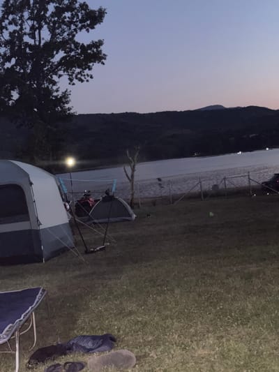 The image depicts a campsite at dusk near a body of water, possibly a lake or reservoir, in Golyama Zhelyazna, Bulgaria. A large, multi-person tent is prominently featured in the foreground to the left, with its entrance flap open. To the right of this tent is a smaller, dome-shaped tent. A bright, circular light source hangs from a pole between the two tents, illuminating the immediate camping area. In the background, beyond a wire fence, the water stretches out, reflecting the dim twilight sky. Hills or mountains form a dark silhouette against the horizon, with a few distant lights visible. The terrain is grassy and appears to be slightly uneven. On the ground in the very foreground, to the left, part of a blue camping chair is visible. Scattered on the grass are dark-colored objects that could be bags or clothing, and a pair of flip-flops. A dark-colored vehicle is partially visible to the right of the fence. The overall atmosphere is serene and suggests the end of a day spent outdoors.