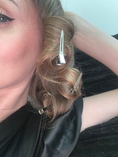 A close-up, overhead shot shows a person's hair being styled with silver clips in Lynnwood, Washington, United States of America. The person, whose face is partially visible on the left side of the frame, has light brown hair with blonde highlights. Their left eye is made up with black eyeliner. The hair is curled and sectioned with multiple silver clips holding the waves in place. The person is wearing a black garment, likely a satin robe, which is draped over their shoulder and chest. The background is a textured dark surface, possibly a blanket or sofa, and a light blue wall. The lighting suggests an indoor setting with natural light coming from the side.