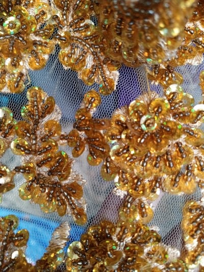 This close-up image showcases a segment of a luxurious textile, featuring intricate gold embellishments on a fine, sheer white mesh fabric base. The primary subjects are the detailed embroidery, sequins, and beads forming a repeating floral or leafy pattern.
Small, circular gold-toned sequins are densely applied, many exhibiting an iridescent quality that reflects subtle hues of green, blue, and pink under the bright lighting. These sequins are arranged in clusters to form petal-like shapes and foliage. Interspersed with the sequins are numerous tiny, elongated seed beads, primarily in a dark brown or deep gold color, which define outlines and add textural depth, particularly along the veins of leaf motifs and within the centers of the floral patterns. Additional gold-colored thread embroidery contributes to the raised, textured appearance of the designs.
Through the transparent mesh, a softly blurred background is visible, revealing patches of blue and purple, suggesting the embellished fabric is laid over another colored material or surface. The lighting is bright, enhancing the sparkle of the sequins and highlighting the three-dimensional quality of the beads and embroidery. The intricate craftsmanship and rich materials evoke a sense of elegance and opulence, characteristic of decorative textiles or traditional attire found in regions such as Houaykhang, Laos.
