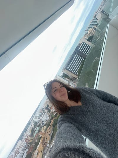A young Caucasian female is depicted in a selfie photograph taken from an elevated position, likely a high-rise balcony, overlooking the city of Les Meravelles, Spain. The female, centered in the lower right quadrant, has medium-length brown hair, a slight smile, and is wearing sunglasses pushed up onto her head. She is dressed in a textured, dark grey V-neck sweater. Her right arm is extended, indicating she is holding the camera device.

The immediate foreground features a portion of a white ceiling or overhang and a glass balcony barrier. Beyond the barrier, the city of Les Meravelles stretches into the distance under an overcast sky. Numerous buildings with varying architectural styles and roof colors, primarily light earth tones, are visible. Further in the background, a row of taller, more modern buildings with darker facades and prominent windows lines what appears to be a coastal area. A large body of water, consistent with the Mediterranean Sea, is visible on the horizon. The overall perspective is tilted, suggesting the photo was taken at an angle.