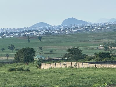 The image depicts a vast, hazy landscape dominated by rolling green hills and a distant settlement. In the foreground, a fence made of concrete posts and panels runs horizontally across the frame. Beyond the fence, the terrain is covered in lush green grass, with scattered trees. A prominent, large tree with dense foliage stands to the left of the frame, near a small green-roofed building.

Further in the background, a sprawling urban area is visible, characterized by a dense collection of houses with white roofs, interspersed with trees. The houses appear to be low-rise structures. Behind the settlement, a range of majestic blue-hued mountains rises, their peaks softened by the atmospheric haze. The sky is a pale, muted blue, suggesting either early morning or late afternoon light. The overall impression is one of a rural or semi-rural setting at the edge of a populated area, likely Gidan Waziri, Nigeria, given the context provided. No people are visible in the image.