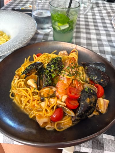A black plate of spaghetti with seafood is centered on a black and white checkered tablecloth. The pasta is topped with mussels, clams, shrimp, cherry tomatoes, and chopped parsley. To the left of the plate is a white bowl of plain spaghetti. To the right of the plate is a clear glass filled with ice, mint, and what appears to be a Mojito cocktail. In the background, another glass of water can be seen. The image suggests a meal at a restaurant in Finale Ligure, Italy.