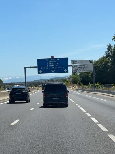 The image shows a highway scene with a bright blue sky. In the foreground, two dark-colored SUVs are driving on the road. The SUV on the left has a license plate that reads "GJ 918 AZ". The SUV on the right has a license plate that reads "HC 794 AQ". In the background, there are tall green trees and distant mountains. Overhead, there are two large highway signs. The sign on the left, marked with "A 43", lists destinations: "TURIN-MILAN", "GENEVE", "GRENOBLE", "CHAMBERY", and "L'ISLE D'ABEAU-CENTRE". The sign on the right indicates "N 6" and lists destinations: "VILLEFONTAINE", "LA VERPILLIERE", "VAULX-MILIEU", and "L'ISLE D'ABEAU-Parc Technologique". There are also speed limit signs visible on the side of the road. The overall scene depicts a typical highway environment with directional signage.