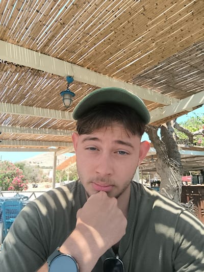 The image shows a young man with light skin, brown hair, and blue eyes, wearing a green baseball cap and a green t-shirt. He is resting his chin on his hand, with his elbow on a table. A smartwatch is visible on his left wrist. The man is looking directly at the camera with a neutral expression.

He is sitting outdoors under a thatched roof canopy. Sunlight filters through the canopy, casting striped shadows on the man and his surroundings. In the background, there are blue chairs and tables, suggesting a cafe or restaurant setting. Further back, there are flowering bushes with pink blossoms, trees with twisted branches, and a distant, hilly landscape under a clear blue sky. The overall impression is of a warm, sunny day in a Mediterranean location.

The scene suggests a leisurely moment, possibly a break from a vacation. The presence of the canopy and outdoor seating implies a seaside or tourist area. The Municipality of Chalki, Greece is mentioned as the location context. There is no visible text in the image.