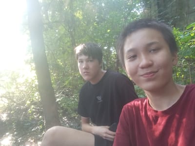 Two young individuals, likely teenagers, are positioned outdoors in a wooded area, with dense foliage and trees forming the background. The lighting suggests it is daytime, with sunlight streaming through the trees, creating a bright, possibly slightly overexposed, atmosphere. One individual, positioned closer to the camera on the right, has short dark hair and is wearing a red t-shirt. Their face is turned towards the camera, and they are smiling. The other individual, on the left, has darker hair and is wearing a black t-shirt with a small white logo. They are looking towards the camera with a neutral expression. One of the individuals is holding a dark object, possibly a phone, in their hands. The environment appears natural and uncultivated, with uneven ground and a significant amount of undergrowth. There is no indication of the location being in the city of Belbulak, Kazakhstan.