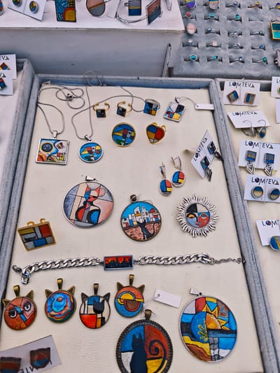 The image shows a display of handmade jewelry for sale, likely at a market or craft fair. The jewelry pieces, many with a painted or enameled finish, are laid out in several trays lined with a light-colored material. The designs are colorful and appear to be inspired by modern art, with geometric shapes, abstract representations of faces, cats, owls, and cityscapes. The brand "LOMTEVA" appears on several of the jewelry display cards. The setting is unknown and there is no confirmation to the location in the city of Uglich, Russia.