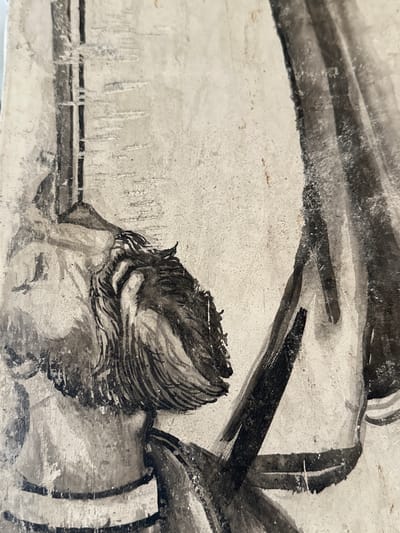 The image shows a detail of a mural painting, likely depicting a historical or religious scene. The style is a fresco, characterized by its muted color palette of grays, blacks, and whites, with some hints of earth tones. The texture of the wall is visible, showing imperfections and the granular nature of the plaster.

In the foreground, the head and shoulders of a figure are depicted. The figure has dark, textured hair, rendered with loose brushstrokes. The face is partially obscured, but the profile suggests a downward gaze. The figure appears to be wearing clothing with striped details. To the right of the figure, a dark, draped element, possibly fabric or a garment, is shown with broad, sweeping brushstrokes, creating a sense of volume and movement. Behind the figure, a vertical structure, possibly a pillar or a column, is present, with linear markings that could represent wood grain or a decorative pattern. The background is a light, textured surface, suggestive of a wall.

This mural is located in the former Franciscan monastery of San Miguel Arcángel in Huejotzingo, Puebla, Mexico. The monastery, founded in the 16th century, is renowned for its well-preserved architecture and significant collection of colonial-era murals, some of which are among the oldest in Mexico.