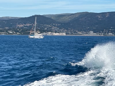 The image captures a wide view of the Mediterranean Sea with a sailboat navigating the choppy blue waters. In the background, the coastal city of Bormes-les-Mimosas, France, is visible, with buildings nestled on the slopes of a verdant, hilly landscape. The shoreline is dotted with structures, and a marina with numerous boats can be seen. The sky is a clear, pale blue, and the sun is shining. The foreground features a wave breaking, creating a spray of white foam. The sailboat, white with a single mast and sail, is positioned to the left of the frame. There are no visible people in the image. The overall scene depicts a tranquil coastal environment with moderate sea conditions.