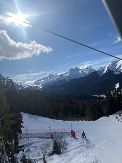 This image displays a sunlit winter scene at a mountainous ski resort near Lunnaya Polyana, Russia. The perspective is from an elevated position, evidenced by a prominent, thick steel cable traversing diagonally from the upper left to the upper right corner of the frame. The sky is clear blue with scattered white cumulus clouds, and a bright sun in the upper left quadrant creates significant glare and lens flare.
In the background, a range of snow-capped mountains rises majestically, their lower slopes and the mid-distance valleys covered by dense, dark evergreen forests. Within one of these forested valleys, a small cluster of buildings, suggesting a village or resort area, is faintly visible.
The foreground and midground feature a wide, groomed, snow-covered ski slope. Approximately seven individuals are observed on the slope, all appearing to be skiing downhill. They are dressed in various colors of winter ski apparel. The ski slope is bordered by dark evergreen trees and equipped with red safety netting held by poles, along with additional orange poles and red triangular markers. Distinct ski tracks are visible on the snow surface, particularly in the foreground.