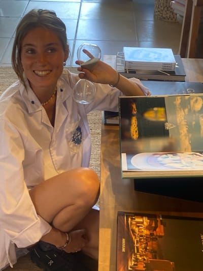 A woman smiles, holding an hourglass in one hand, while sitting near a coffee table stacked with art books. The books feature titles like "Chanel," "Gaudi," and "Assouline." She wears a white button-down shirt. Her legs are bent, with one foot tucked beneath the other. The scene is bathed in soft natural light.