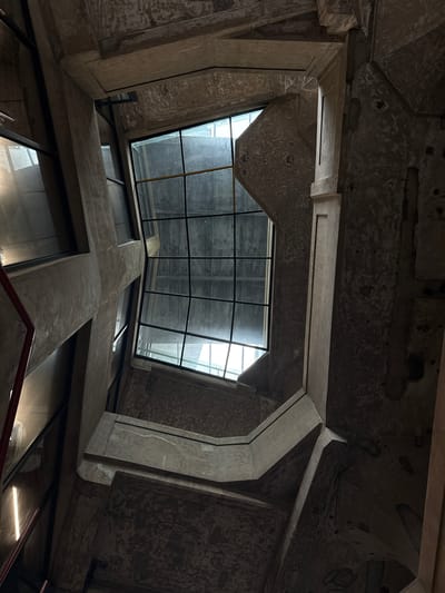 The image presents an upward view of an architectural space located in Hongkou District, China, characterized by a Brutalist style. The space is dominated by exposed concrete elements forming a complex geometric structure, including a cross-shaped formation in the lower portion of the frame. The ceiling consists of a grid of glass panels supported by metal beams, allowing natural light to penetrate the space. The concrete surfaces exhibit signs of aging and weathering. No people are visible. Artificial lighting is present within the space.