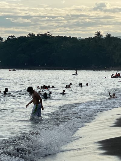 In Dauin, Philippines, a group of people are enjoying the ocean near the shore. The foreground shows a man wading in the shallow water with waves breaking around him, his lower body submerged. He is wearing light-colored shorts with a patterned design. Behind him, a crowd of people are swimming and playing in the water, with some appearing to be in groups. Further out in the sea, there are individuals on paddleboards and a small boat with people. The background features a dense line of trees, likely a tropical forest, bordering the coastline. Above the trees, the sky is overcast with scattered clouds. The overall scene depicts a lively beach day with people engaged in various water activities.