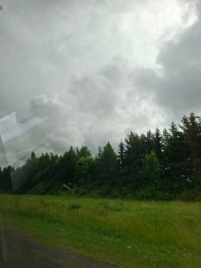 The image depicts a cloudy sky over a dense forest bordering a grassy field. A gravel road is visible in the foreground on the left side of the image. Reflections on what appears to be a window or glass surface distort parts of the scene, particularly in the upper left quadrant, showing geometric shapes and faint landscape elements. The overall impression is a view from inside a moving vehicle on a grey, overcast day. The location is specified as Pokrovka, Russia. There are no people or animals present in the image.