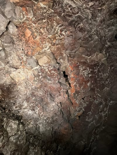 The image captures a detailed view of a textured rock surface, likely within a cave or lava tube, consistent with the volcanic landscape of Haría, Spain. No people or artificial objects are visible.

The scene is dominated by rough, uneven rock formations with a fractured and layered appearance. The primary colors of the rock are dark browns, various shades of gray, and reddish-browns, suggesting volcanic origin. Numerous white and off-white streaks, patches, and irregular patterns are prominent across the entire surface, possibly indicating mineral deposits, flowstone, or lighter veins within the rock. Some areas of the rock also exhibit a soft orange or reddish glow, which may be a result of the mineral composition or specific artificial lighting.

The lighting is localized and uneven, highlighting the intricate textures and variations in the rock while leaving other areas in deep shadow, reinforcing the impression of an artificially illuminated, dark interior space. The intricate patterns and varying shades create a geological and almost abstract composition. There is no visible text in the image.