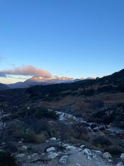The image depicts a mountain landscape, likely taken at sunrise or sunset. The sky is a gradient of blue. Snow-capped peaks are visible, some illuminated by a warm, golden light. There's a cloud formation with similar warm tones near the base of the mountains. The foreground shows a rocky, vegetation-covered slope. A small stream or waterfall is visible in the rock terrain. The photograph's location is given as the city of Unknown, Argentina.
