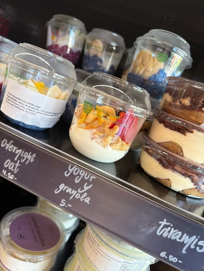 This media file shows a close-up view of a refrigerated display case containing various pre-packaged desserts and breakfast items. The primary subjects are the food items in clear plastic containers, such as tiramisu, chia pudding with spirulina, overnight oats, yogurt with granola, and Labneh de Cabra. The setting is a retail environment, likely a grocery store or market, as suggested by the product labels and display case.
The items are stacked and arranged, with some containers showing layers of ingredients like cake, cream, fruit, and grains. Labels are visible on some of the containers, providing information about the product name, ingredients, and nutritional value. There are also price tags and descriptive signs accompanying the products. One sign, in particular, has handwritten text indicating "tiramis" for 6.50, "yogur y grayola" for 5, and "Overnight oat" for 4.50.
The lighting appears to be artificial, typical of an indoor display. There are no people visible in the image, focusing the attention entirely on the products. The overall impression is one of a well-stocked and organized food display, highlighting convenience and variety. The location context of Santa Eulària des Riu, Spain, suggests a European market or store. The image is sharp and in focus, allowing for clear observation of the food items and text.