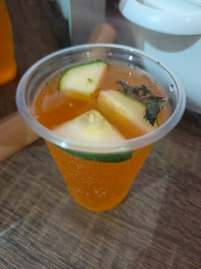 A clear plastic cup filled with an orange beverage sits on a wooden surface. Slices of cucumber and a few sprigs of mint are submerged in the drink. The cup has a green rim near the top. In the background, a blurry white object, possibly a dispenser or appliance, is visible. The overall lighting is dim, with some highlights on the cup and beverage. The scene does not provide any visual cues to determine its location in Kawo, Nigeria.