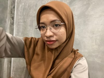 A young woman wearing a brown hijab and glasses poses for a selfie in front of a gray, textured wall. She is looking at the camera with her mouth slightly open, revealing her lips. She is wearing a light gray sweater with a ribbed texture. The lighting is dim, creating a shadow on the left side of her face. The image appears to have been taken indoors, possibly in a residential setting or a cafe in Sekar Mawar, Indonesia, though the location cannot be definitively confirmed from the visual evidence alone.