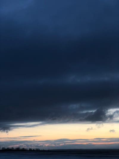 This landscape image, taken in Balakhta, Russia, presents a dramatic sky over what appears to be a large body of water and distant land. The scene is dominated by a stark contrast between dark, brooding clouds and a lighter, warmer horizon.
The left two-thirds of the image are enveloped in heavy, dark blue-grey clouds, which appear dense and potentially indicative of an overcast or stormy weather front. The texture of these clouds varies, with smoother, almost solid dark expanses blending into more textured, slightly lighter formations.
Towards the right side, the sky brightens significantly, transitioning into a band of pale yellow and soft orange hues near the horizon, suggesting either sunrise or sunset. Several thin, dark horizontal lines, resembling power lines or utility wires, are faintly visible stretching across this brighter section of the sky.
Below the horizon, on the far right, a dark, undulating line defines a distant treeline or landmass, appearing as a silhouetted stretch of nature. The area immediately beneath the warm-toned sky, extending to the bottom edge, seems to be a body of water, reflecting some of the ambient light and darkness.
The image captures a moment of powerful atmospheric conditions. There are no people, explicit activities, or visible text in the frame.